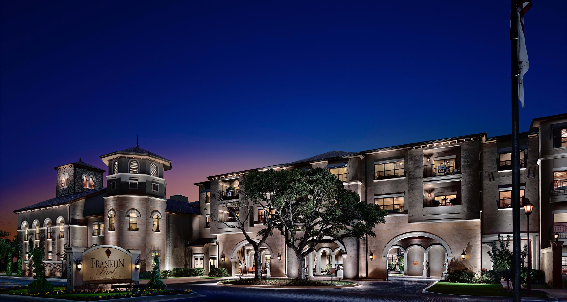Exterior of a Franklin Park Senior Living community at dusk, featuring illuminated stone architecture, arched entrances, and landscaped grounds.