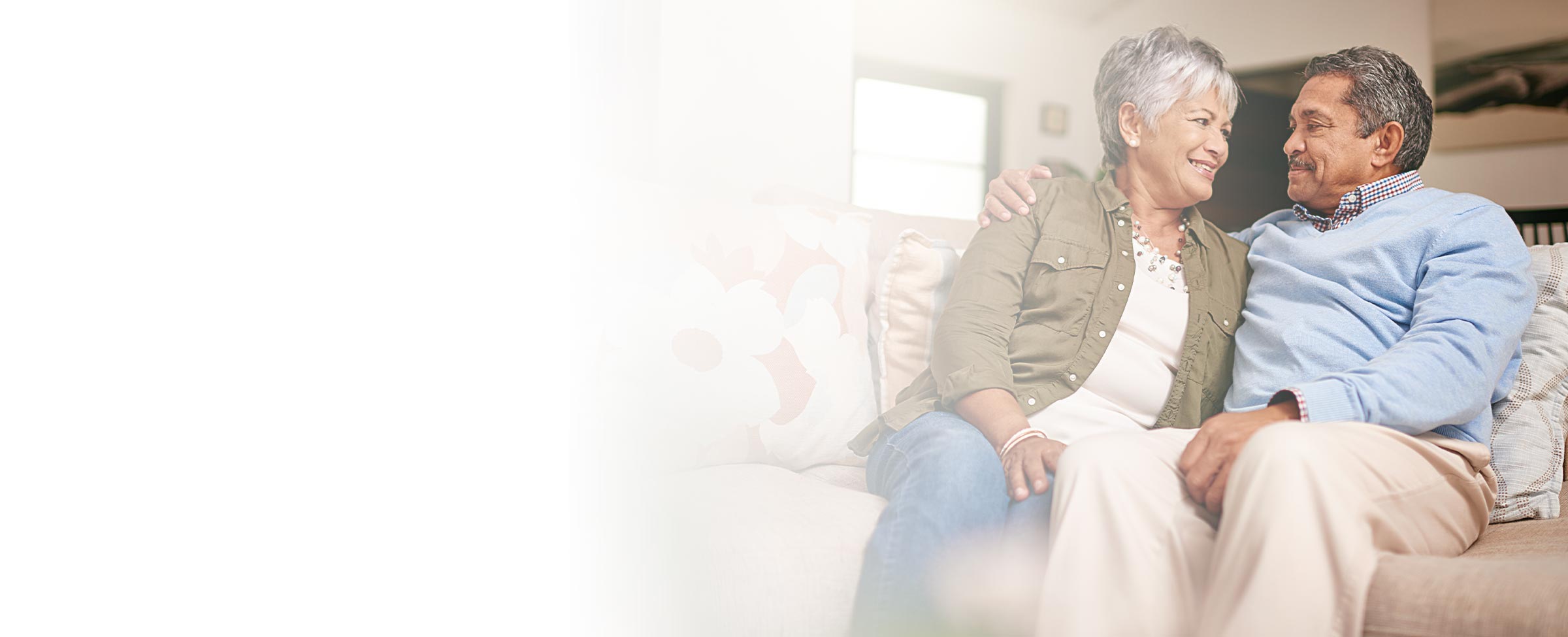 Senior couple sitting on sofa smiling at each other.